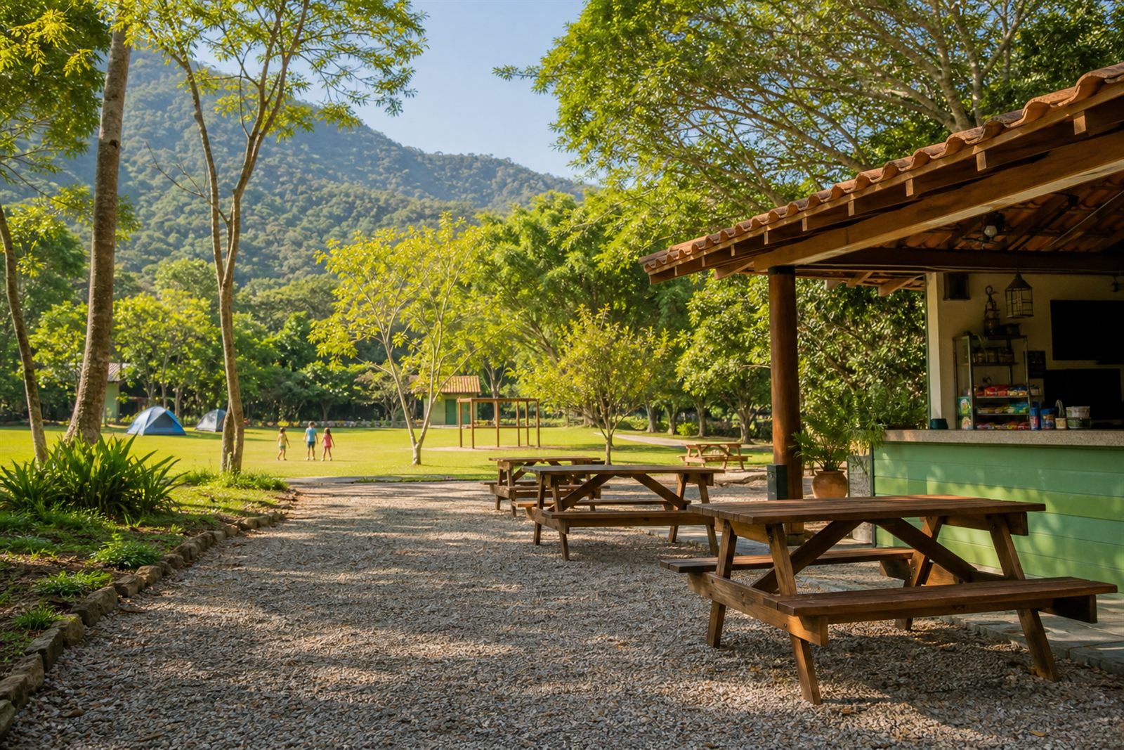 Área de convivência de camping com mesas de madeira, quiosque e gramado com barracas ao fundo